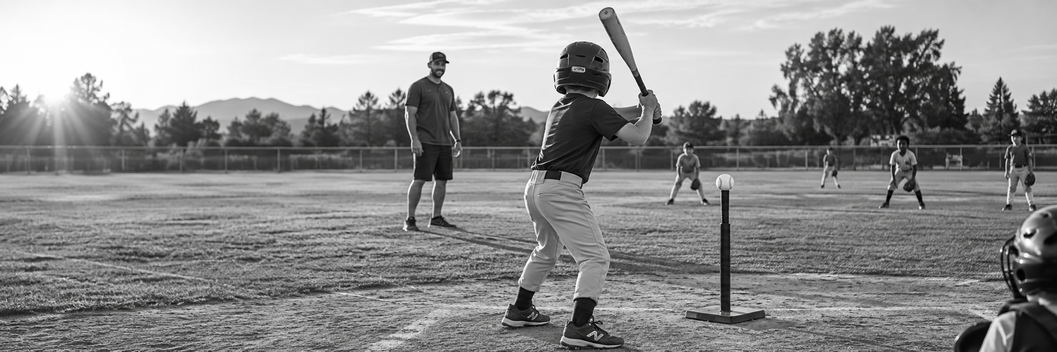 A helmeted young batter preparing to hit a ball from a T-ball tee, with a coach nearby and young fielders ready
